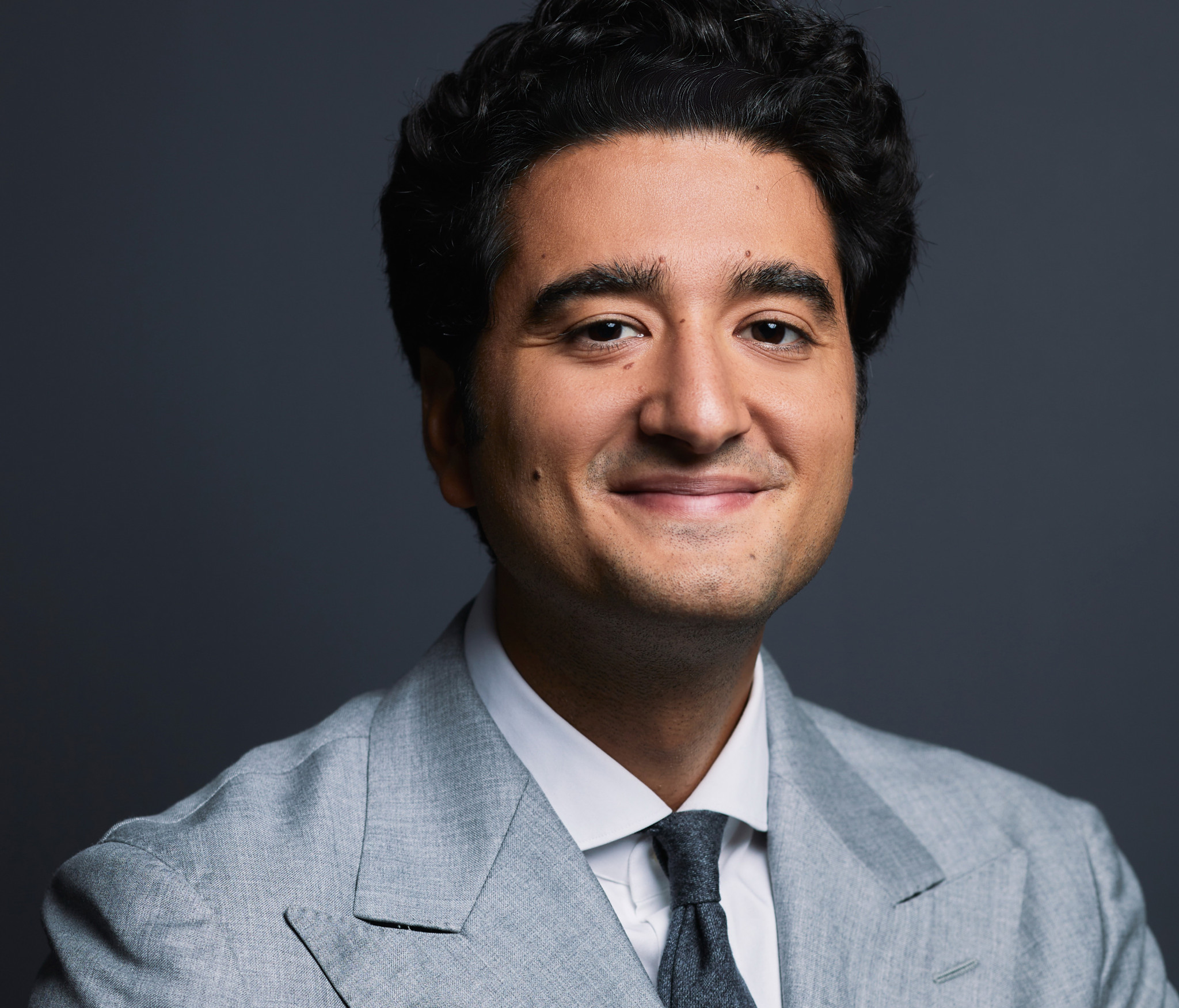 Portrait d’un jeune homme souriant en costume gris clair et cravate grise, sur fond sombre.