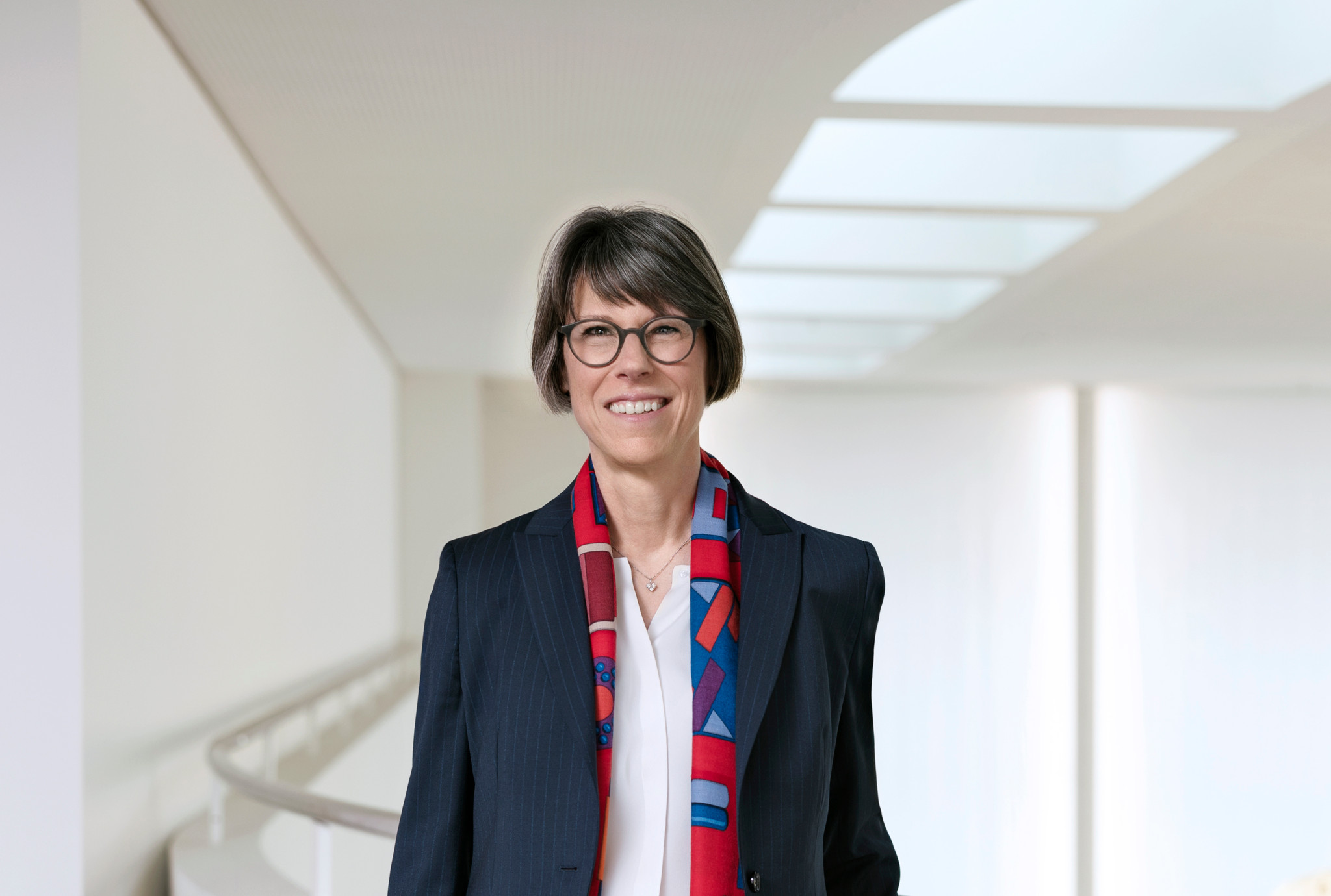 Femme souriante portant un blazer bleu marine et un foulard coloré rouge et bleu, dans un espace lumineux moderne.