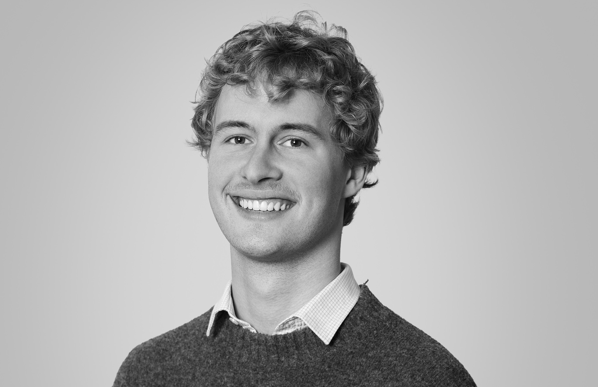 Portrait en noir et blanc d’un jeune homme souriant aux cheveux bouclés, portant un pull en laine sur une chemise à col.