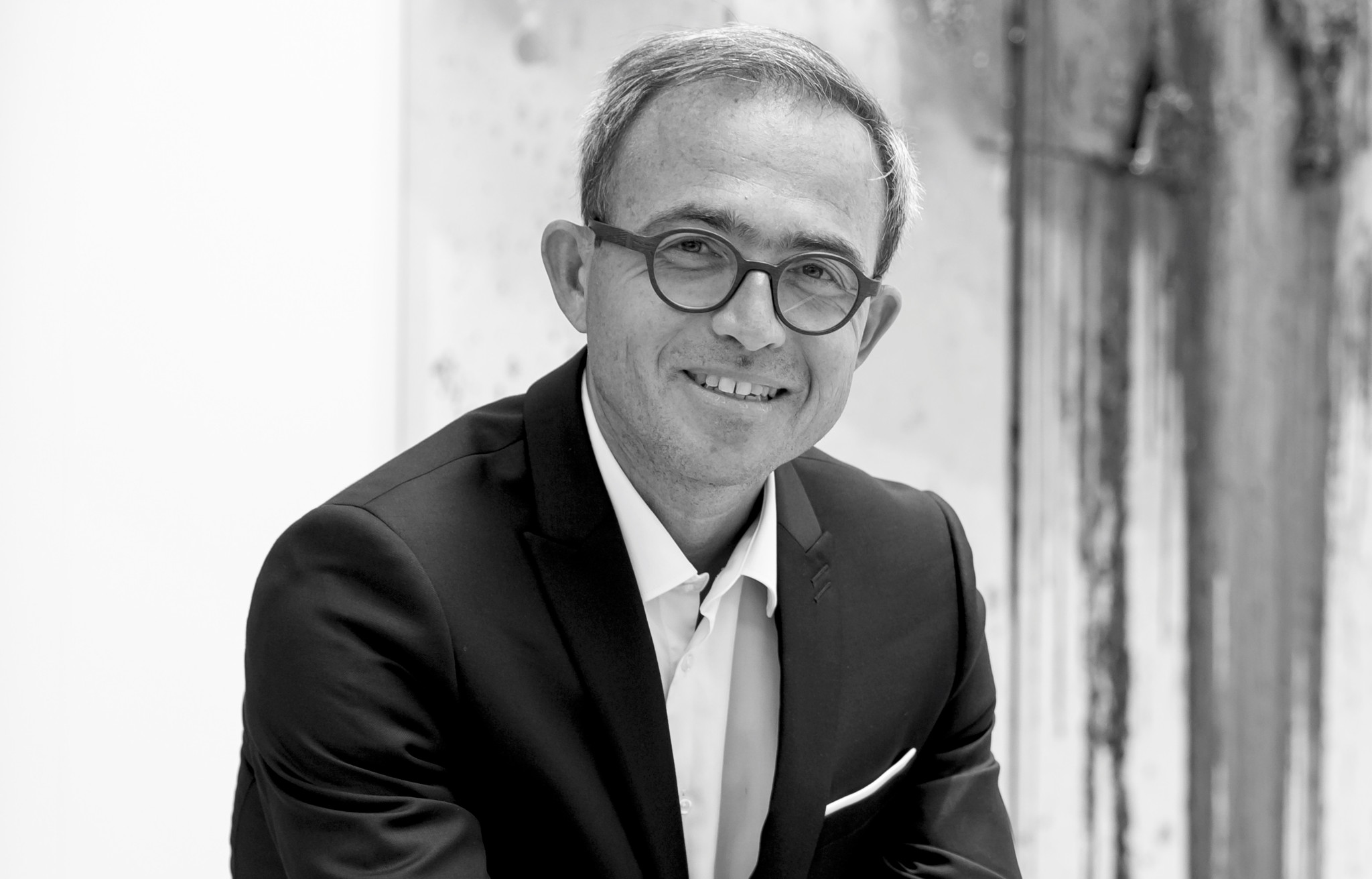 Portrait en noir et blanc d’un homme souriant portant des lunettes rondes et un costume sombre avec une cravate claire.