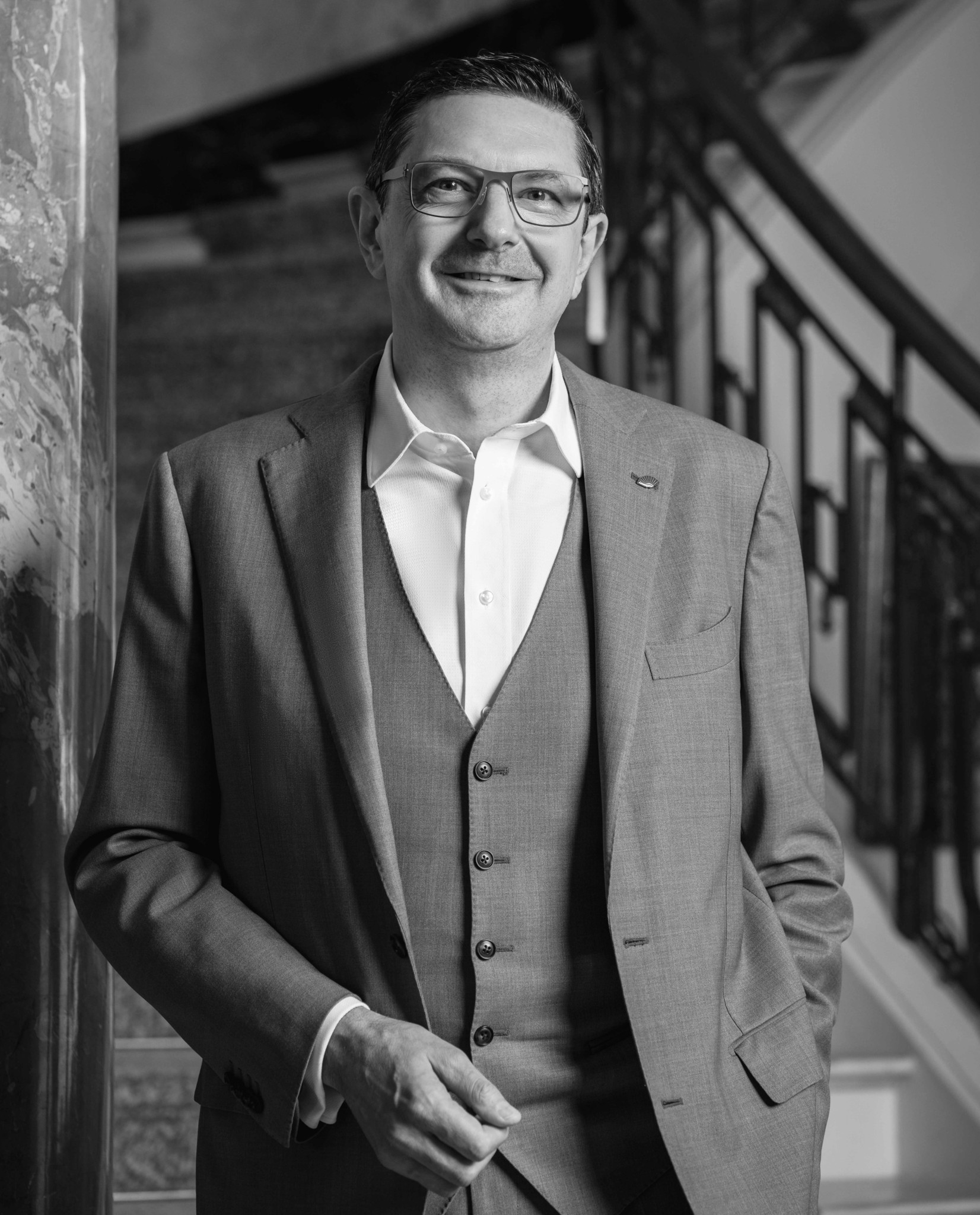 Homme en costume trois pièces gris souriant, portant des lunettes, posant devant un escalier élégant, photo en noir et blanc.