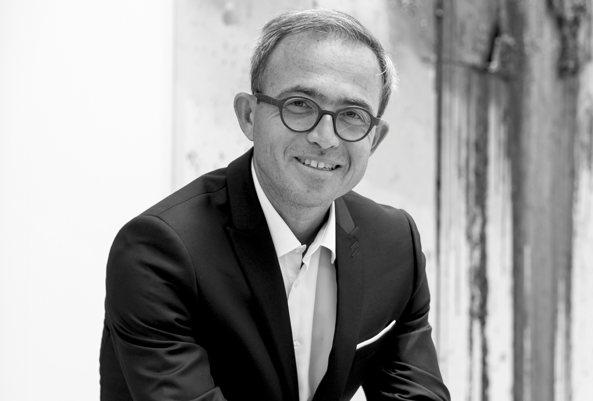 Portrait en noir et blanc d’un homme souriant en costume sombre et lunettes rondes, posant devant un fond texturé.