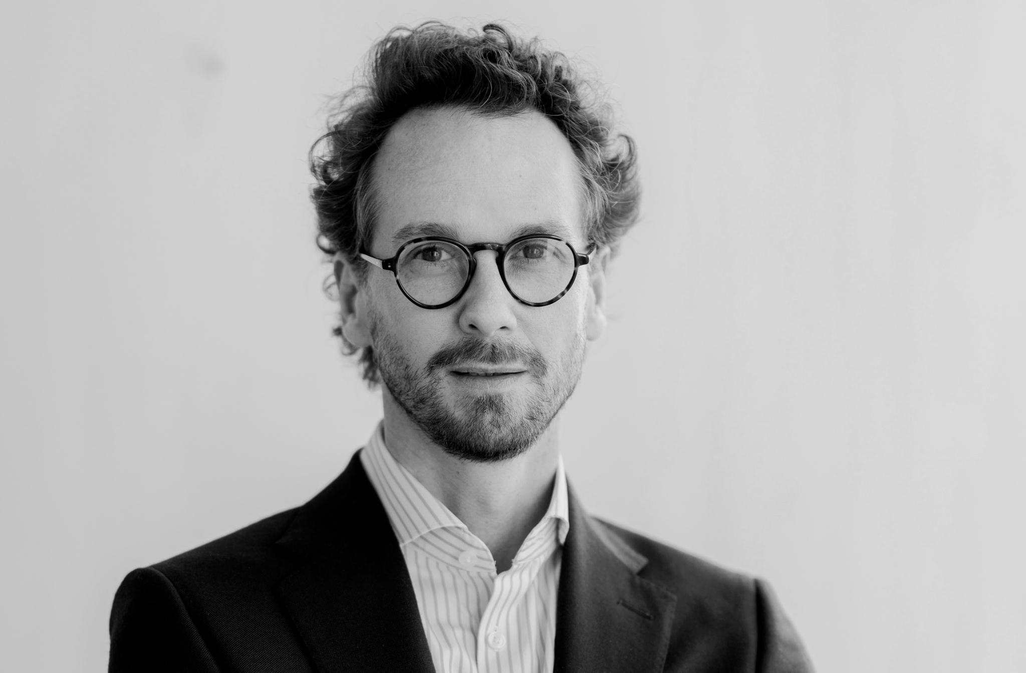 Portrait en noir et blanc d’un homme portant des lunettes rondes, veste sombre et chemise rayée, cheveux bouclés et barbe légère.