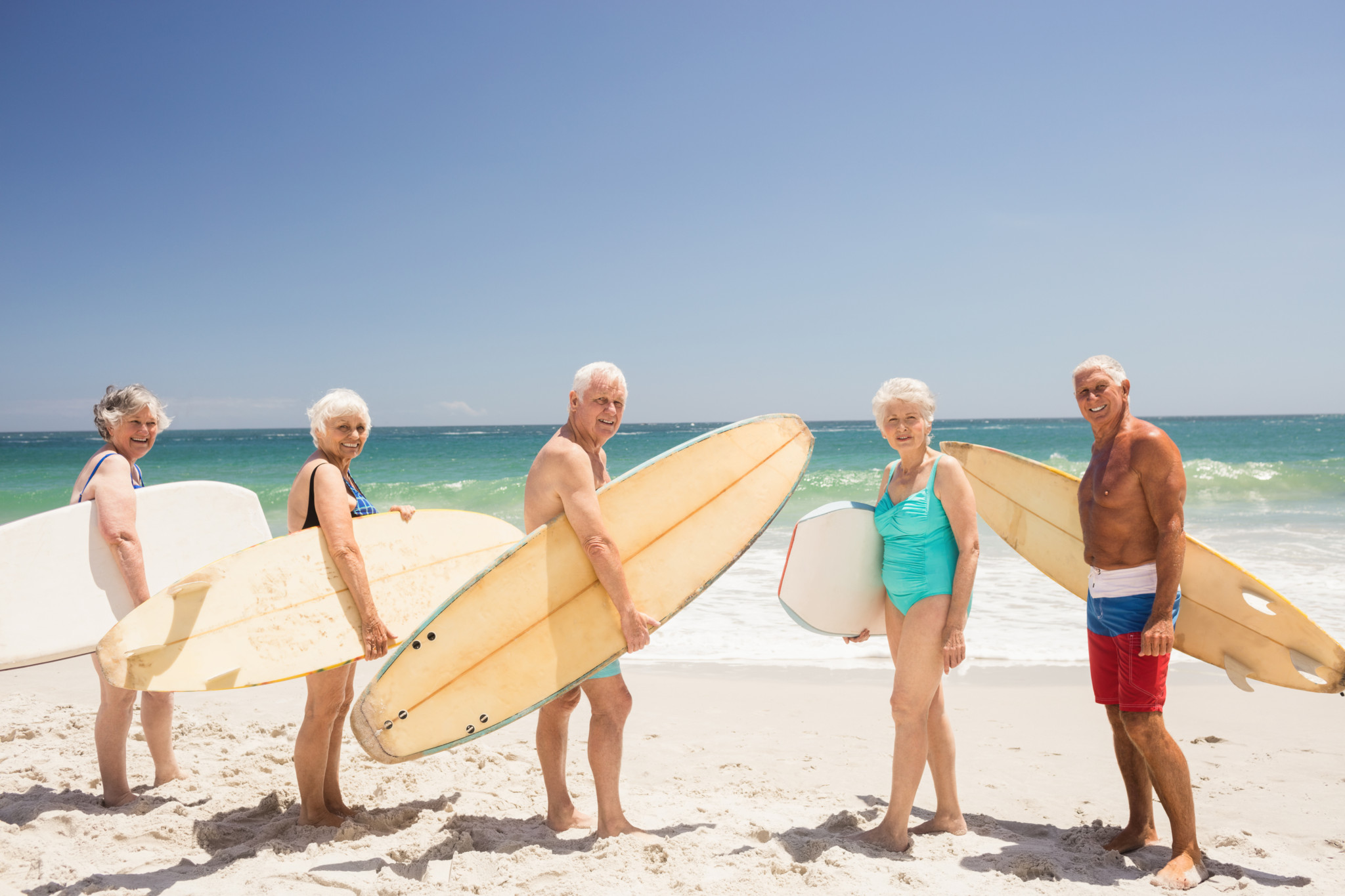 Groupe d’amis seniors tenant des planches de surf sur une plage ensoleillée, souriants face à la caméra