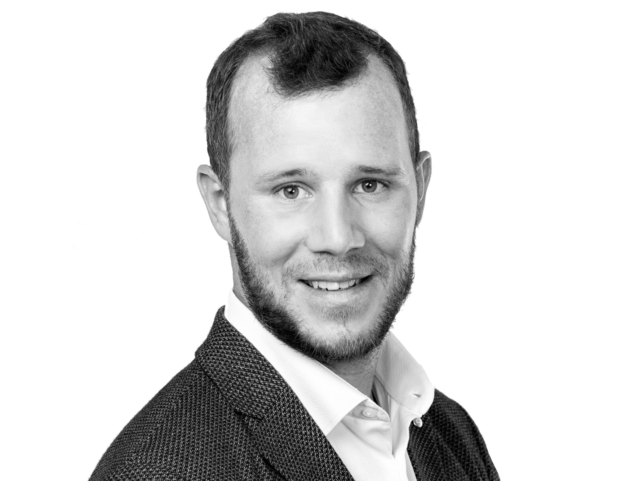 Portrait en noir et blanc d’un homme souriant avec une barbe courte, vêtu d’un blazer et d’une chemise blanche.