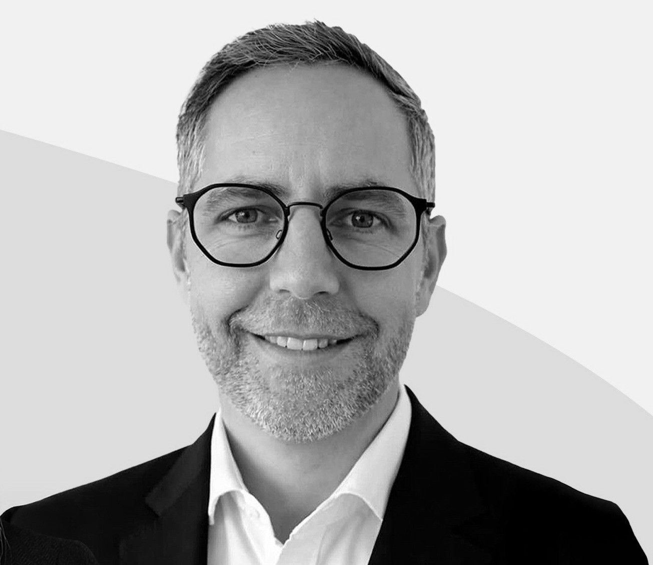 Portrait en noir et blanc d’un homme souriant portant des lunettes rondes et un costume sombre, sur fond clair.