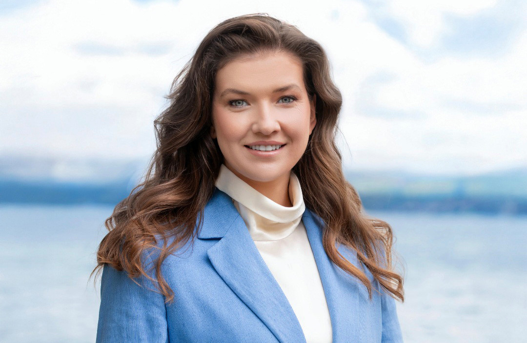Une femme souriante aux longs cheveux bruns, vêtue d’un blazer bleu et d’un pull col roulé crème, posant devant un lac et des montagnes.
