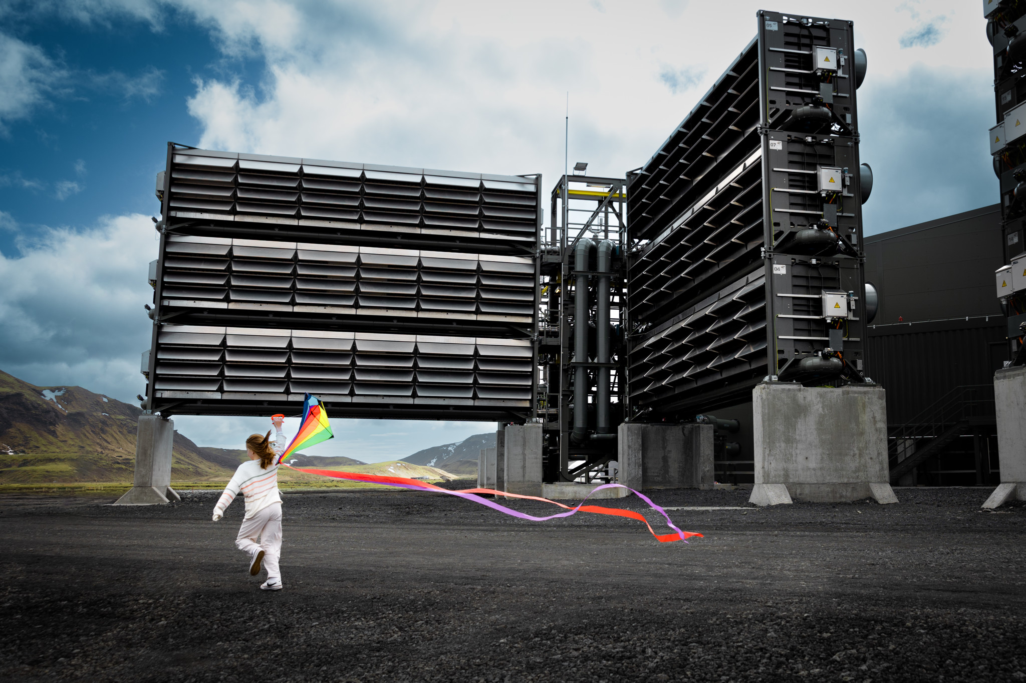 Une enfant court avec un cerf-volant coloré devant des grandes structures industrielles sous un ciel nuageux.