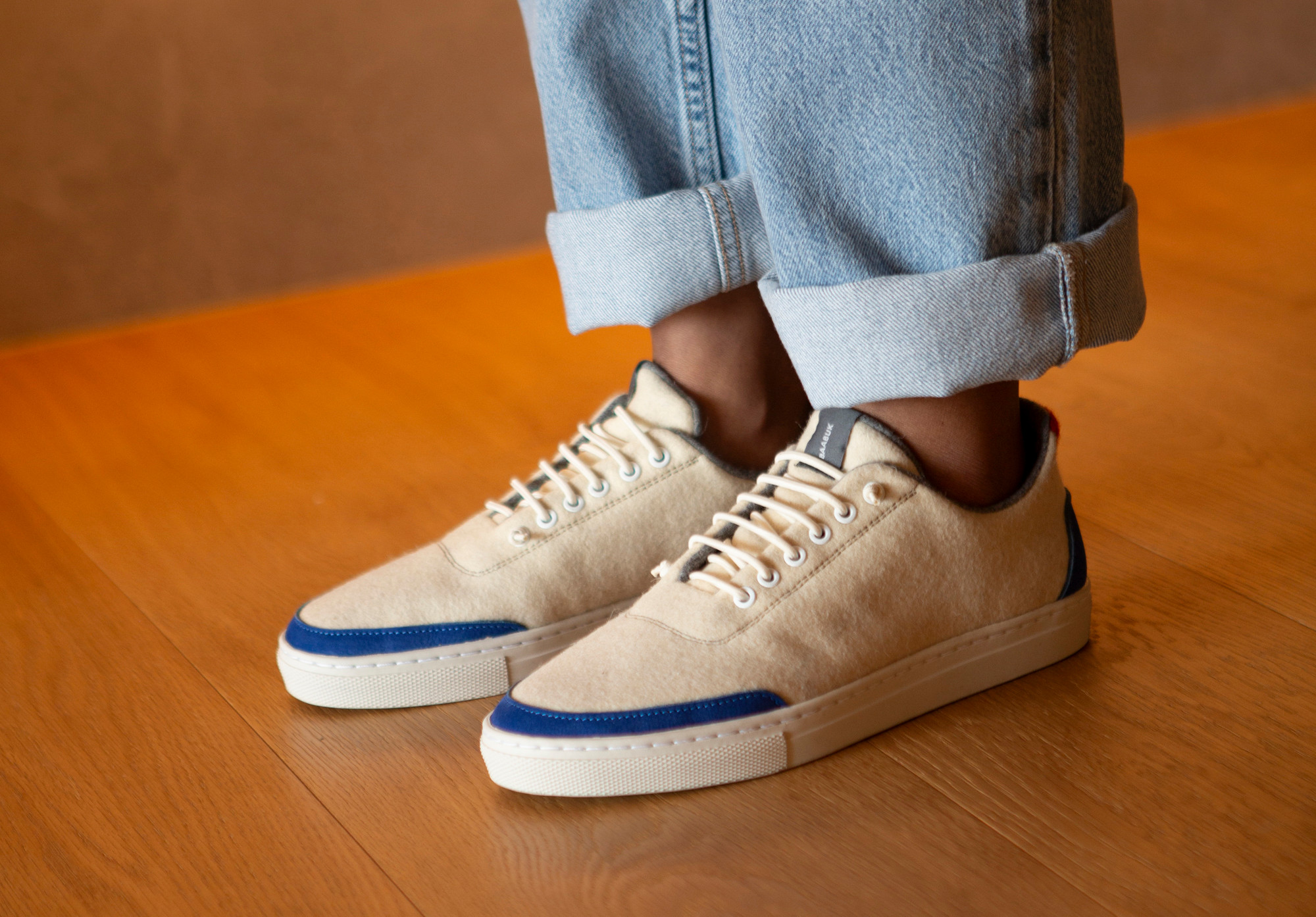 Sneakers beiges avec détails bleus portées avec un jean retroussé, posées sur un parquet en bois