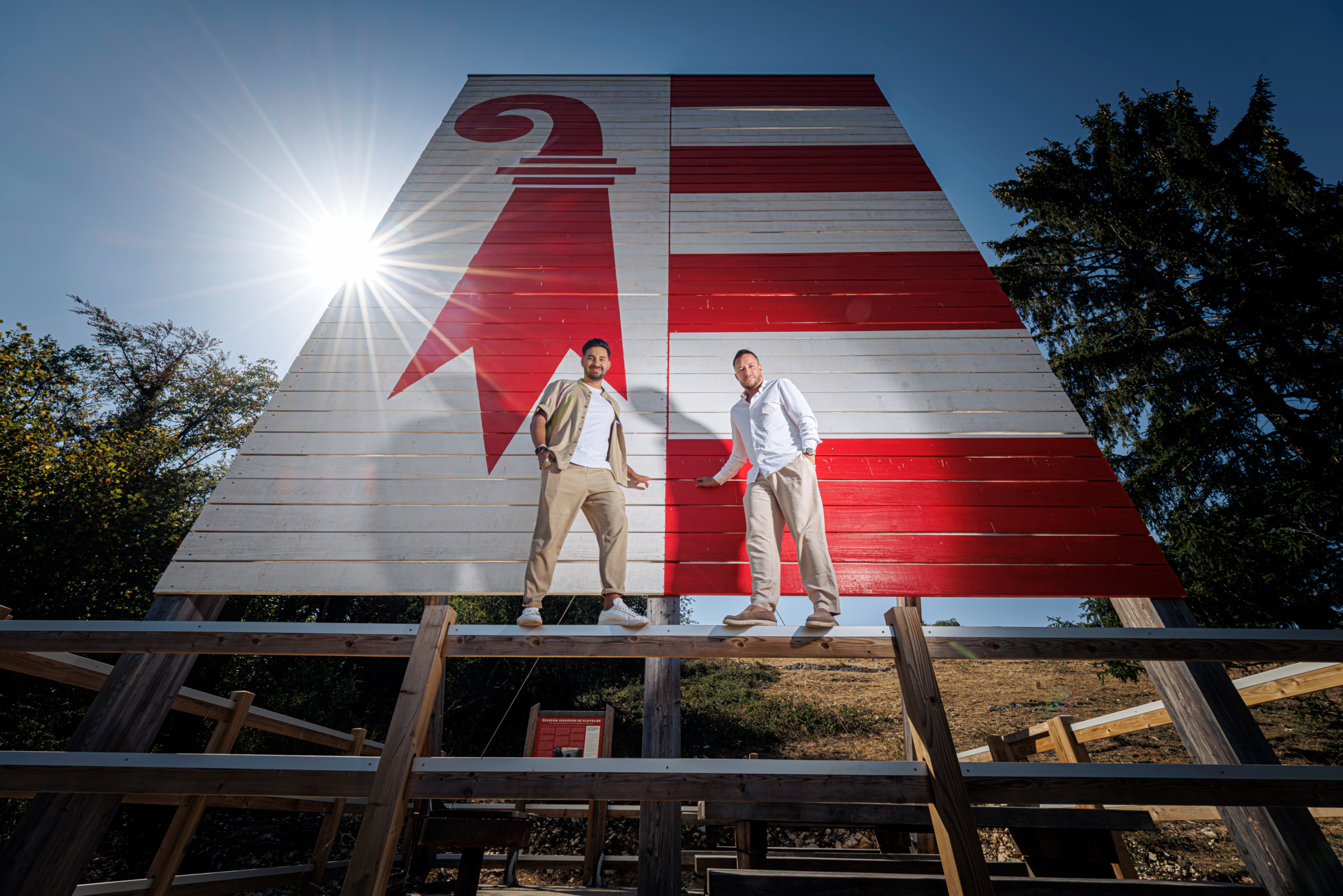Deux hommes debout sur une structure en bois devant une grande fresque murale rouge et blanche représentant un drapeau, sous un soleil éclatant.