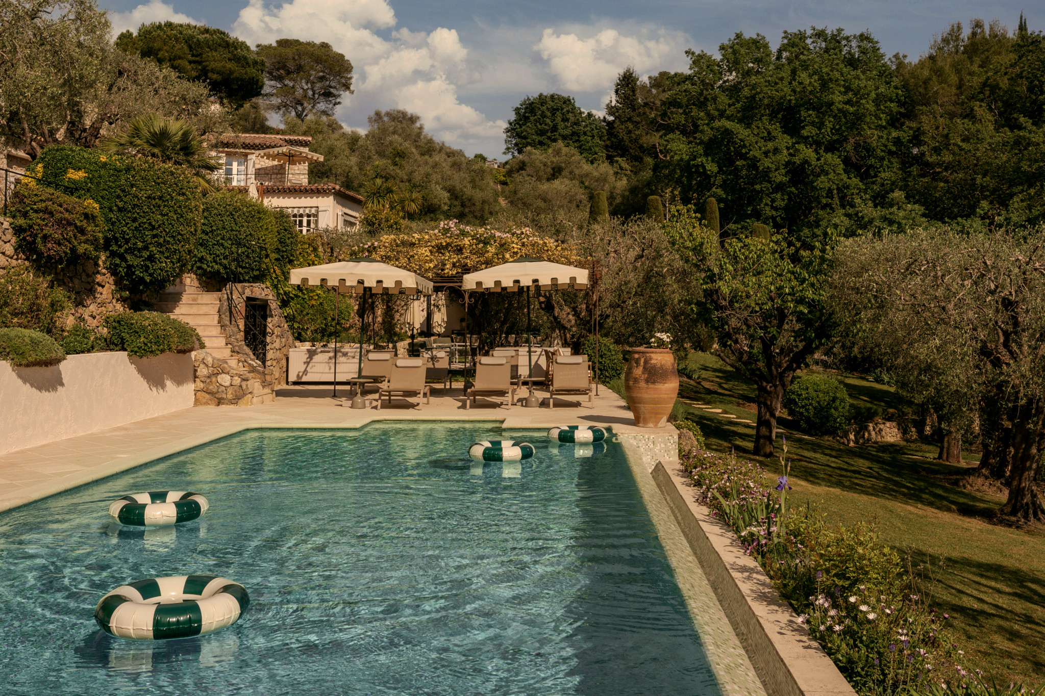 Piscine extérieure luxueuse avec bouées rayées vert et blanc, entourée d’oliviers et d’une villa provençale sous un ciel ensoleillé.