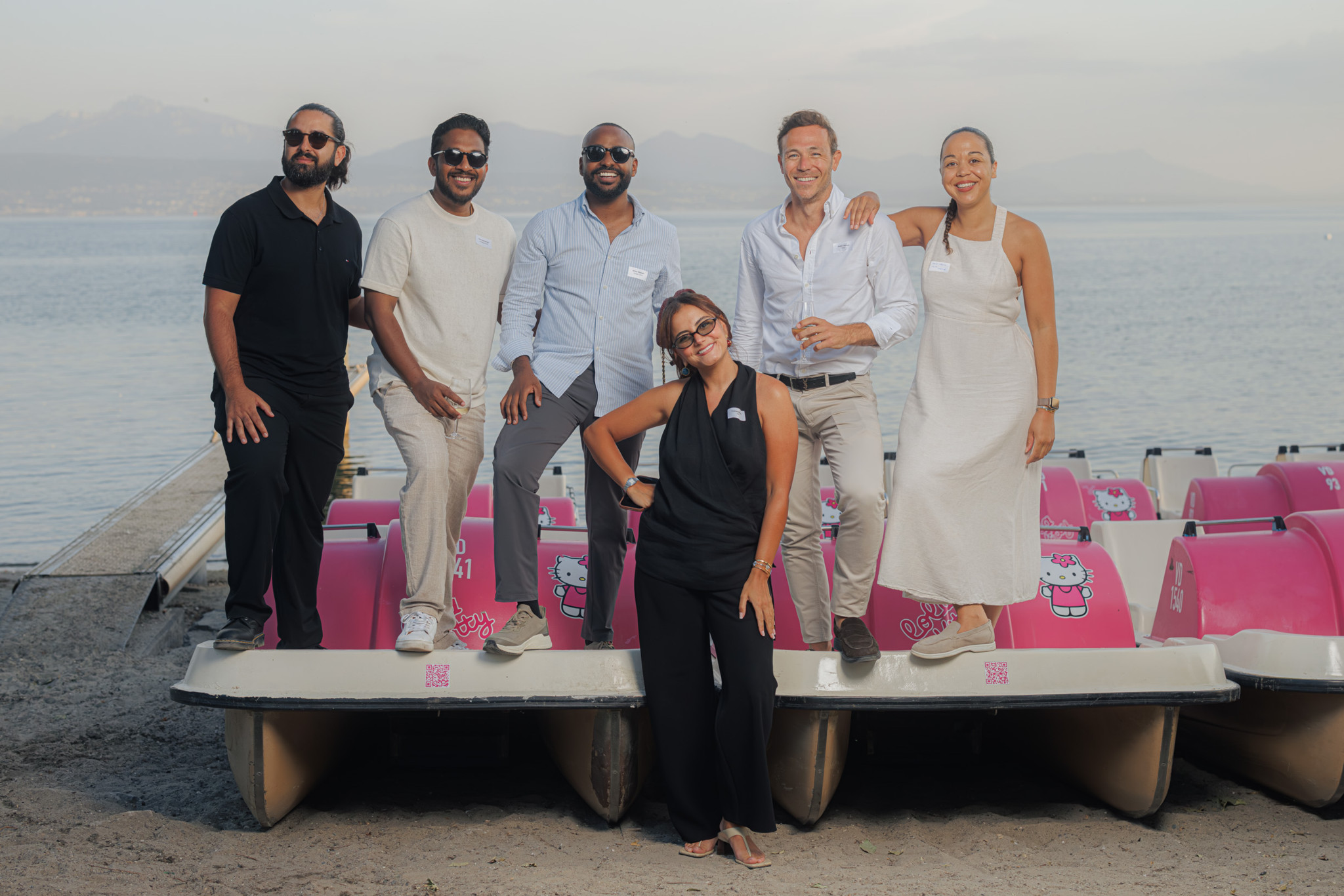 Groupe de six personnes souriantes posant devant des pédalos roses Hello Kitty sur une plage au bord d’un lac.