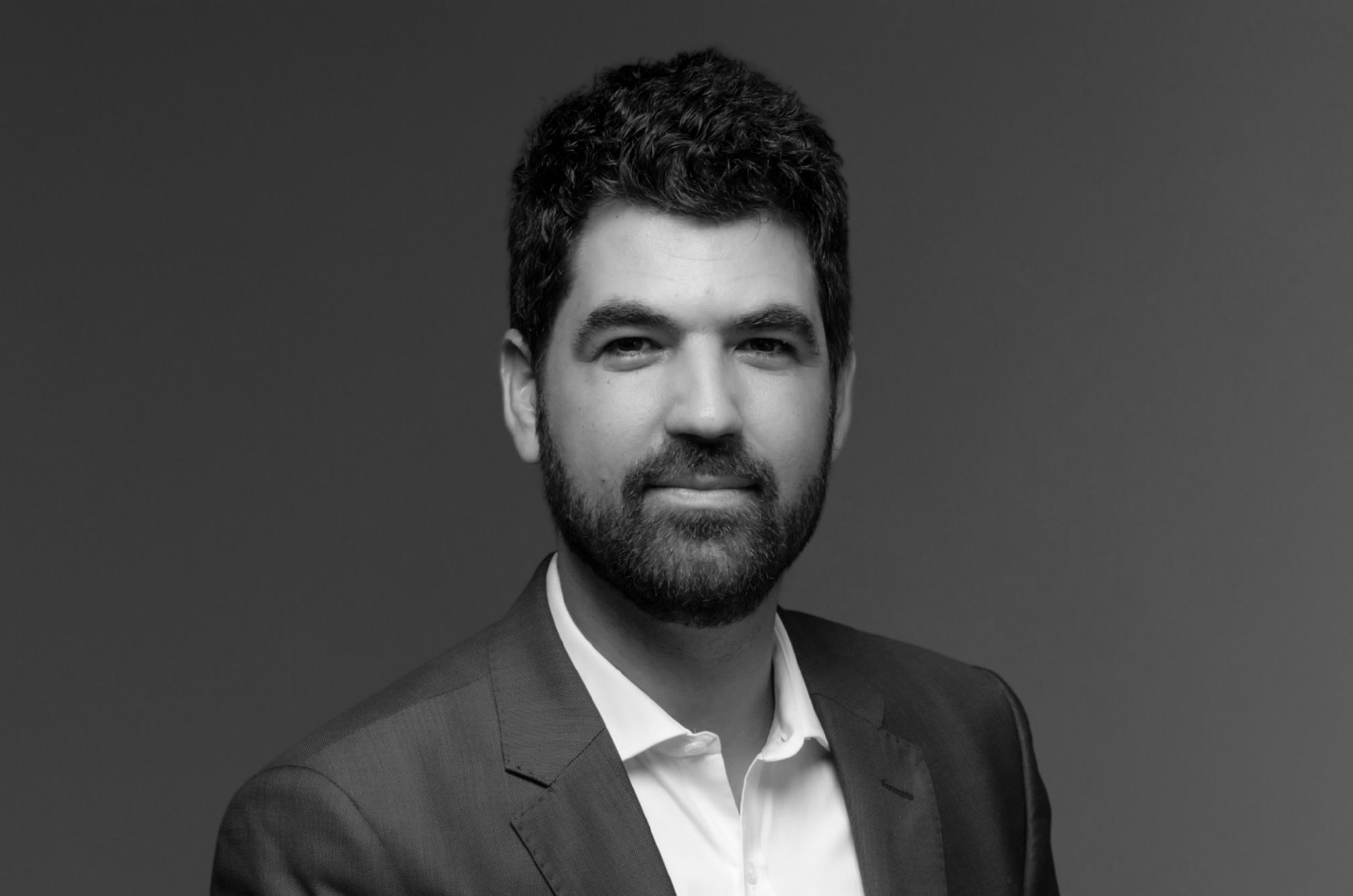 Portrait en noir et blanc d’un homme brun avec une barbe courte, portant un costume sombre et une chemise blanche.