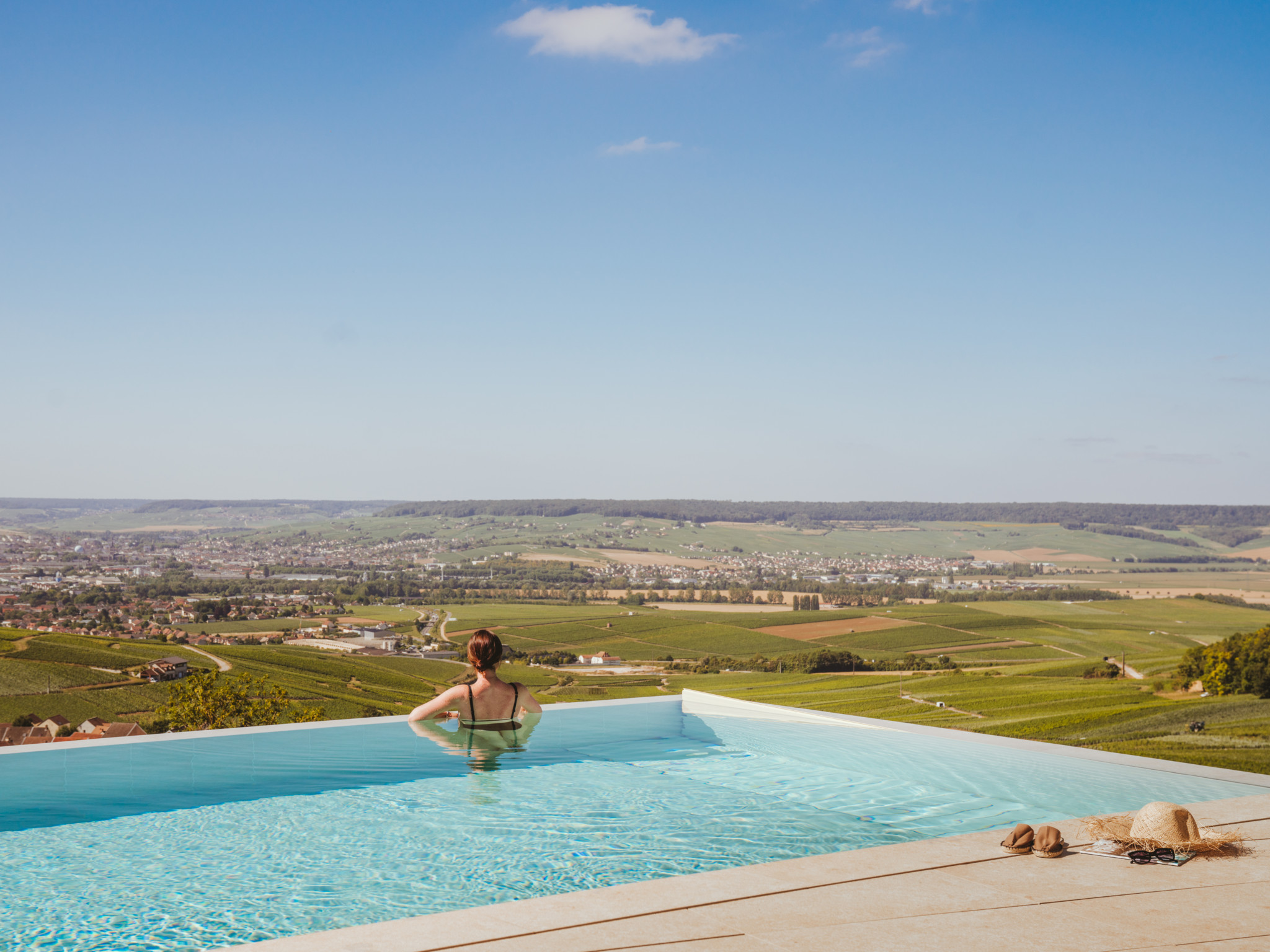 Une femme dans une piscine à débordement contemple un panorama de vignobles et de campagne sous un ciel bleu ensoleillé.