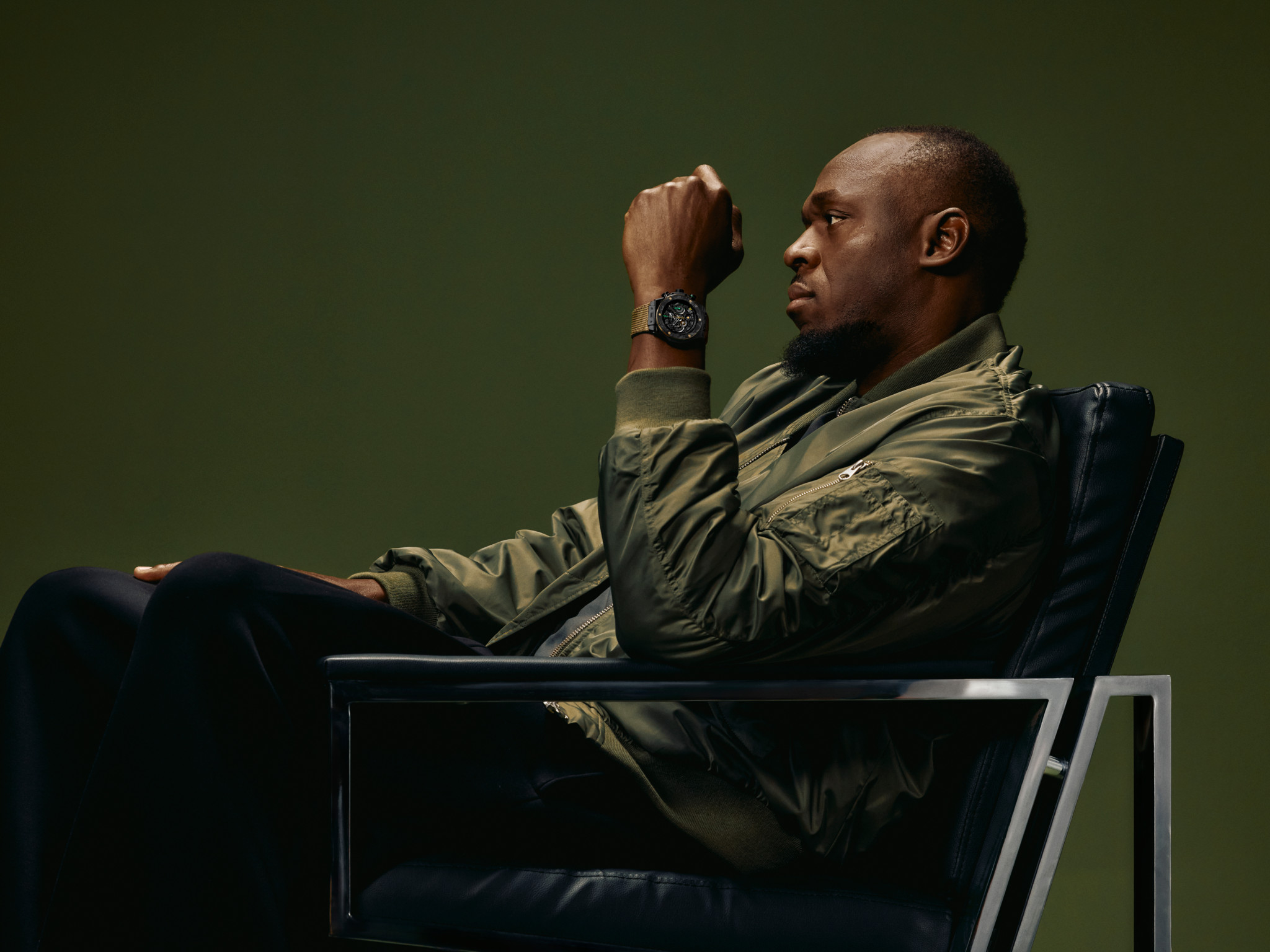 Homme en veste kaki assis dans un fauteuil design, portant une montre noire, sur fond vert sombre
