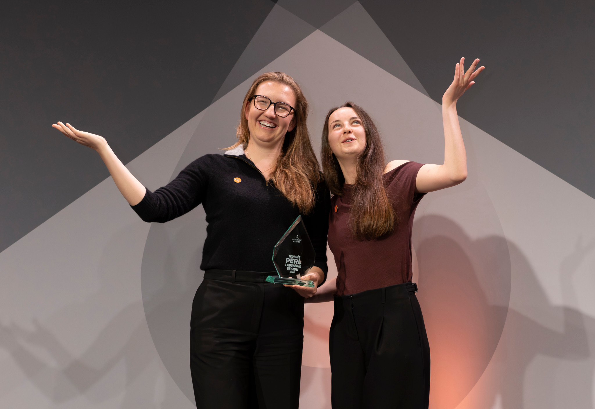 Deux femmes souriantes sur scène tenant un trophée en verre, les bras levés en signe de célébration lors d’une cérémonie de remise de prix.