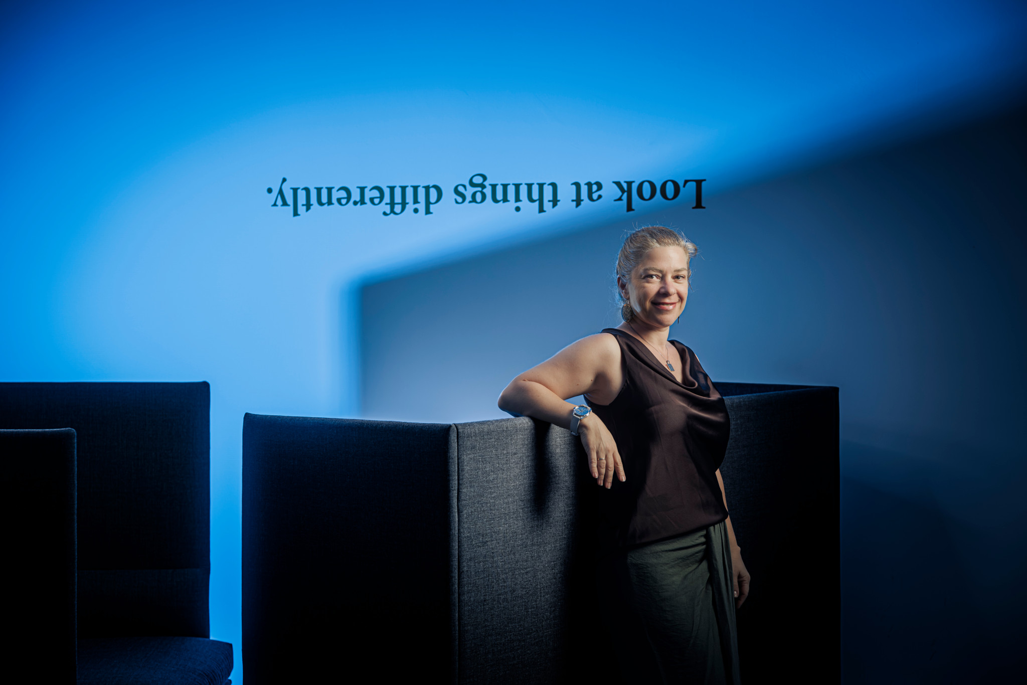 Une femme souriante accoudée à une cloison grise, devant un mur bleu avec l’inscription inversée «Look at things differently.»