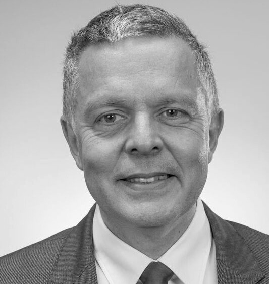 Portrait en noir et blanc d’un homme d’âge mûr aux cheveux courts, portant un costume et une cravate, souriant légèrement.