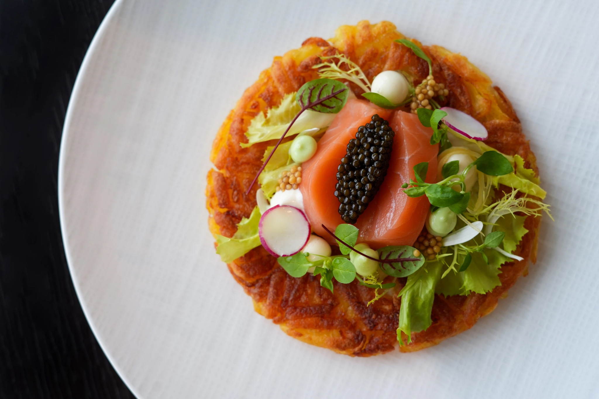 Rösti croustillant garni de saumon fumé, caviar noir, radis, micro-pousses et moutarde, servi sur assiette blanche