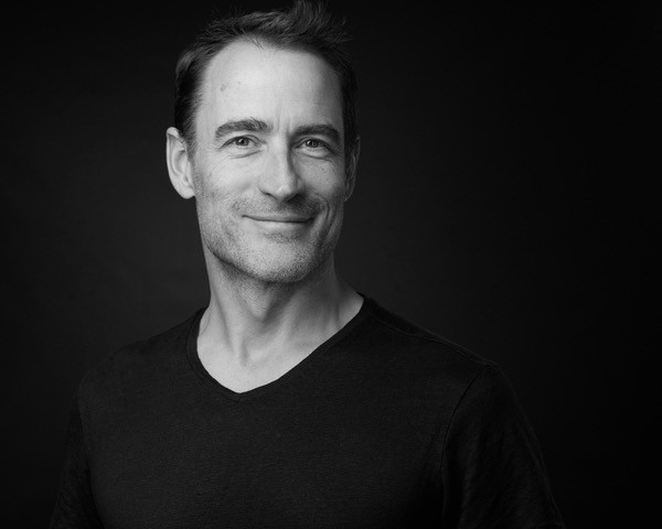 Portrait en noir et blanc d’un homme souriant, vêtu d’un t-shirt noir, sur fond sombre.