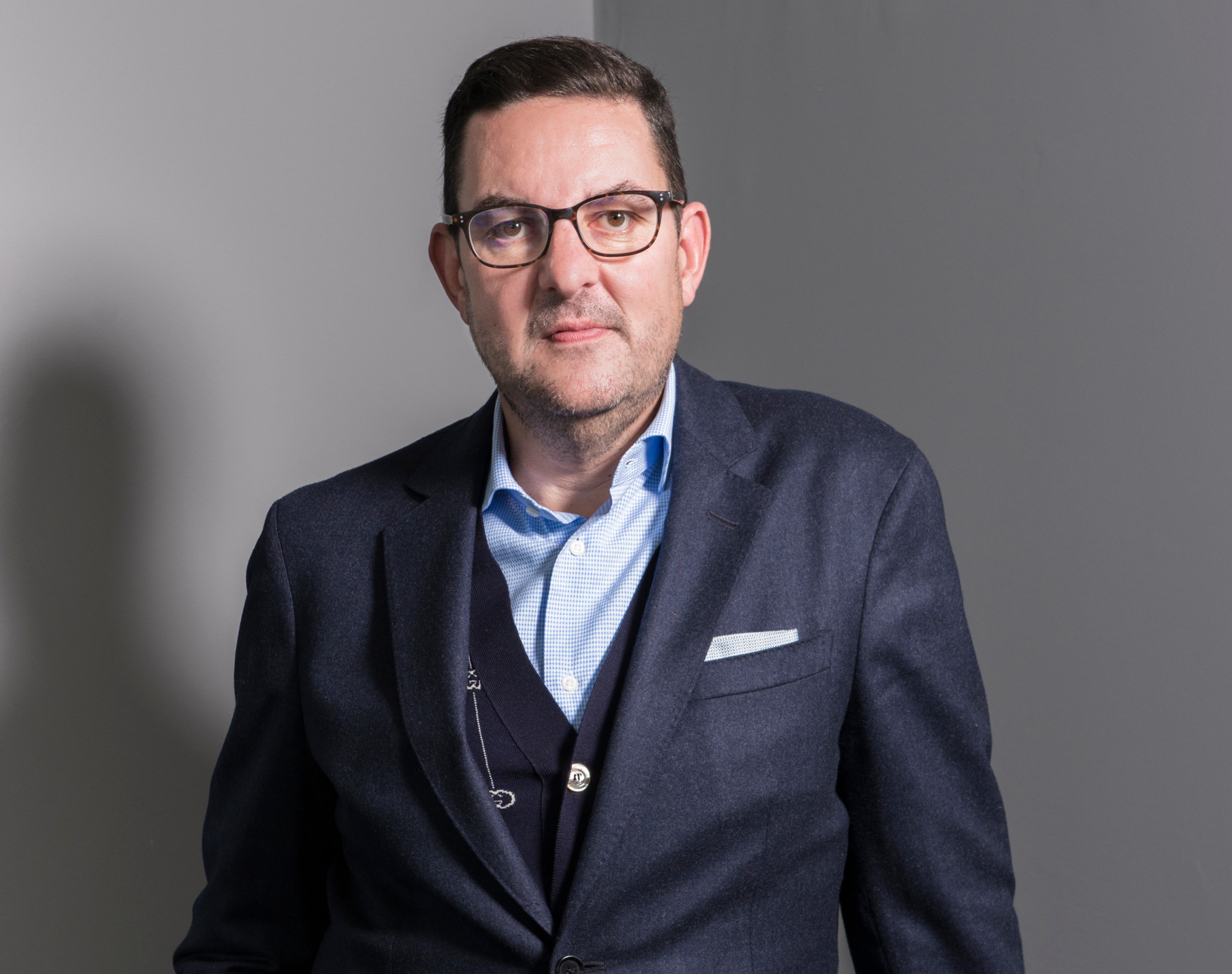 Homme en costume bleu marine et chemise bleue claire, portant des lunettes, posant devant un fond gris neutre.