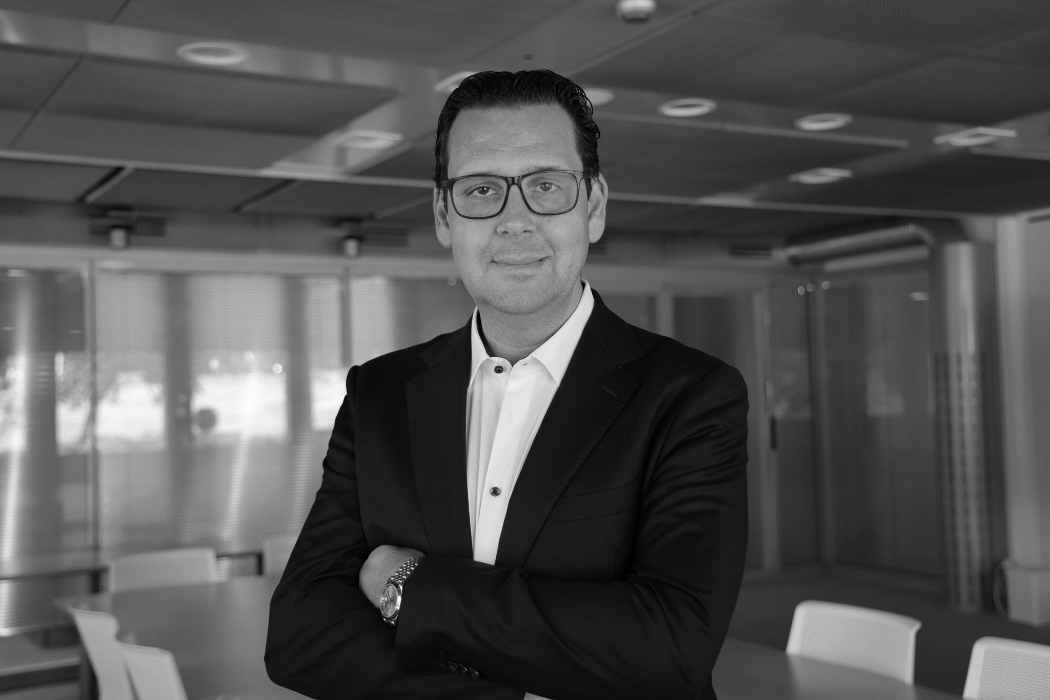 Homme en costume sombre et lunettes, bras croisés, souriant dans un bureau moderne, photo en noir et blanc.