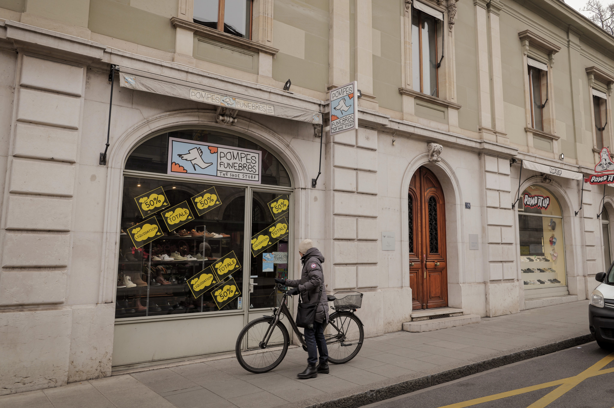 Devanture du magasin de chaussures Pompes Funebres, Rue de la Corraterie à Genève, affichant des pancartes de réduction. Une personne à vélo passe devant.