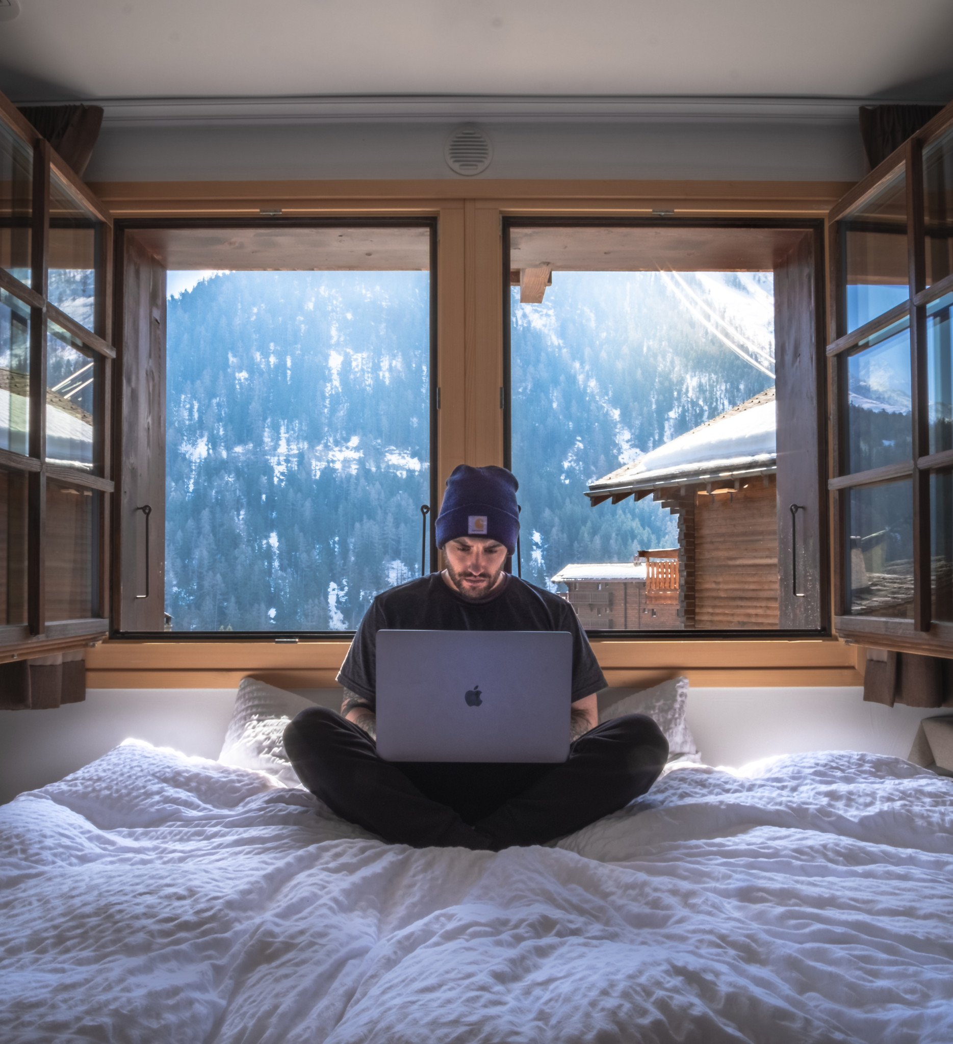 Homme assis sur un lit avec un ordinateur portable dans une chambre avec vue sur des montagnes enneigées à travers une grande fenêtre.