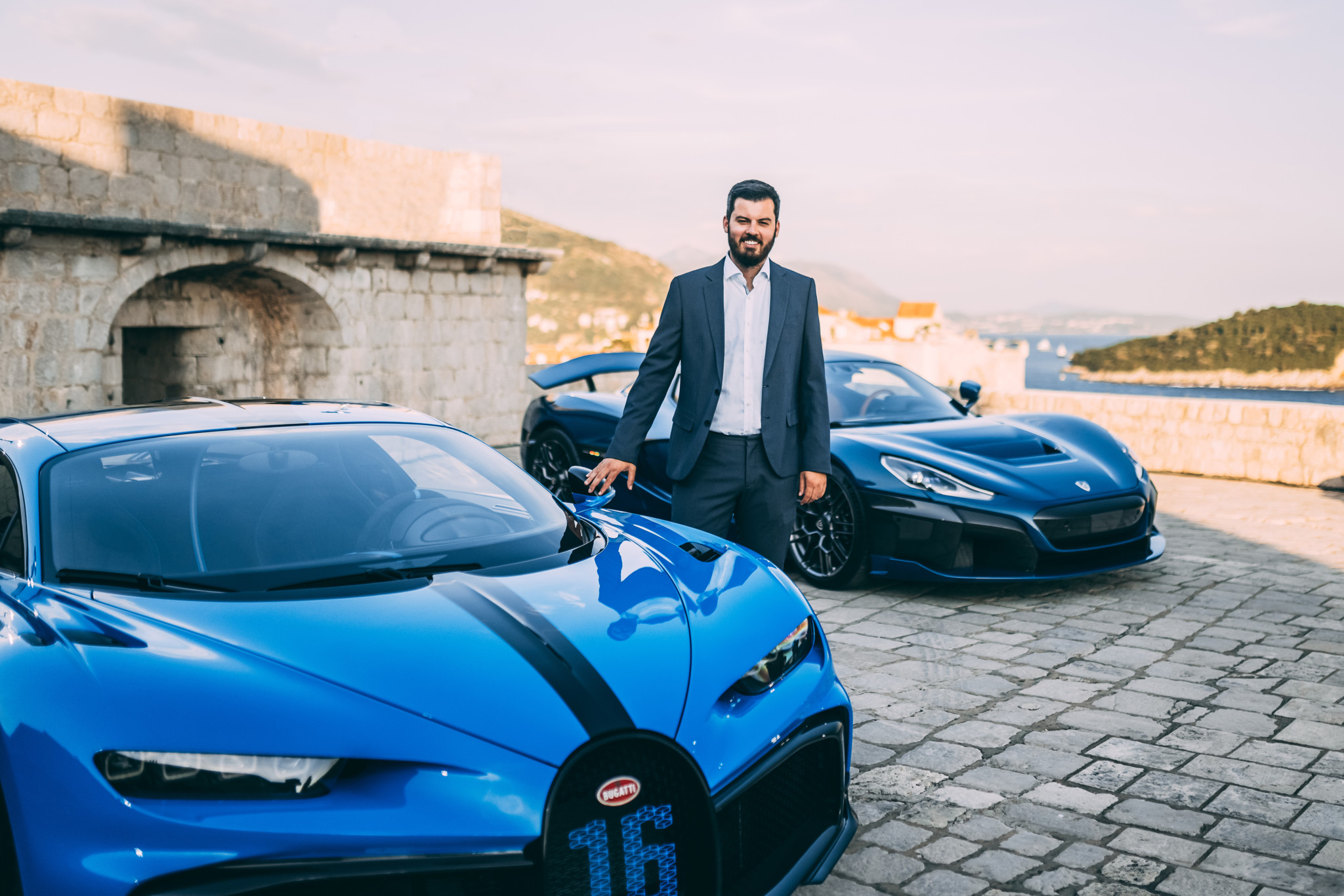 Un homme en costume posé entre deux supercars de luxe, dont une Bugatti bleue, devant un paysage méditerranéen.