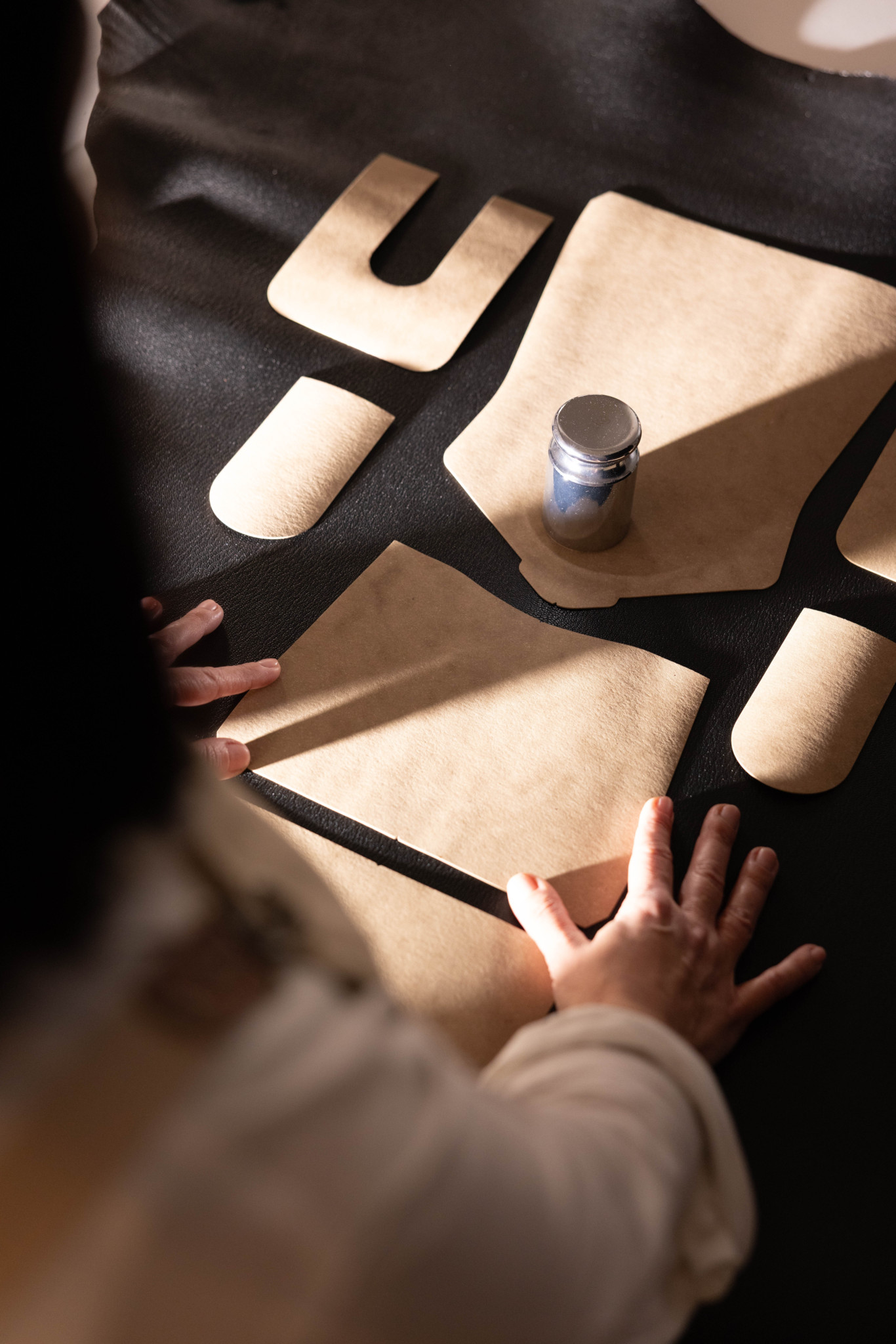 Mains d’une personne manipulant des pièces de cuir beige découpées sur une surface sombre, avec un pot métallique posé à proximité.